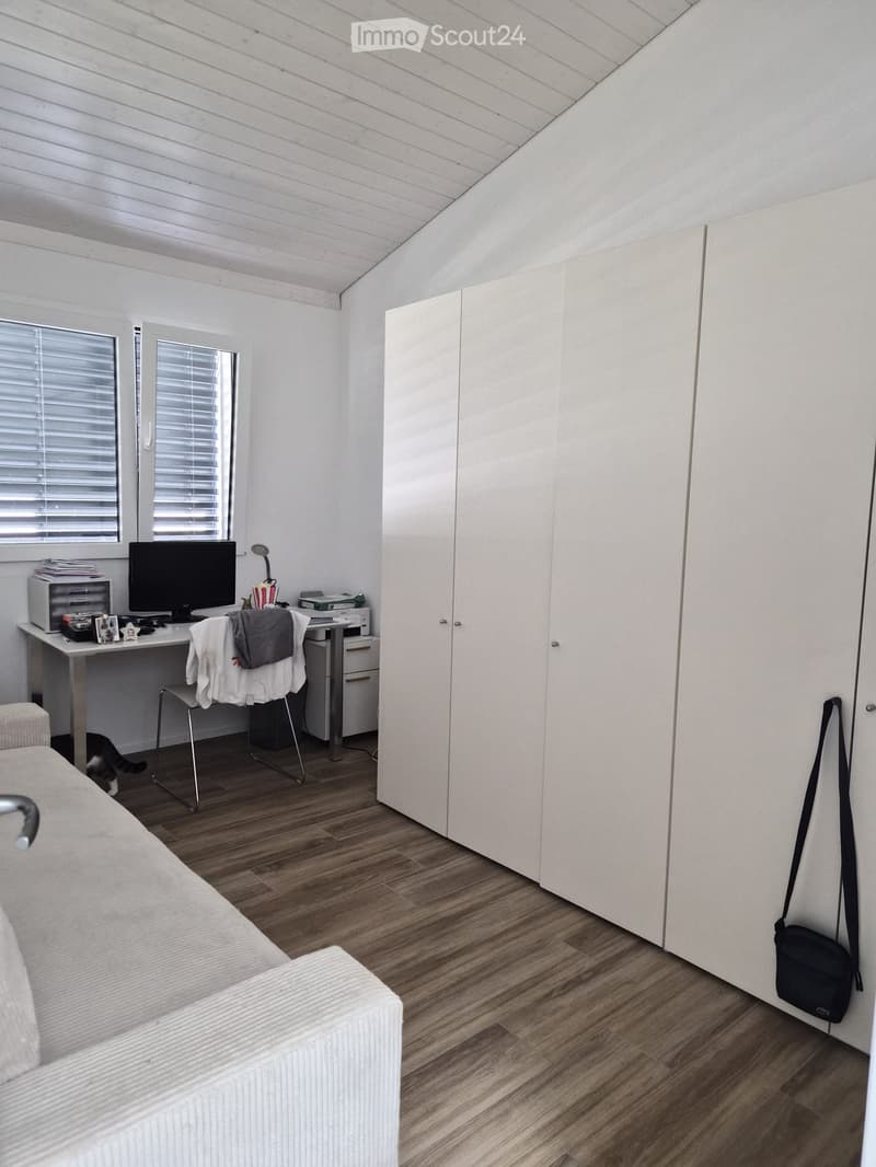 A furnished room with a sofa, a chair, a table with a screen, a cat, and a handbag. The room has wooden flooring and white walls.