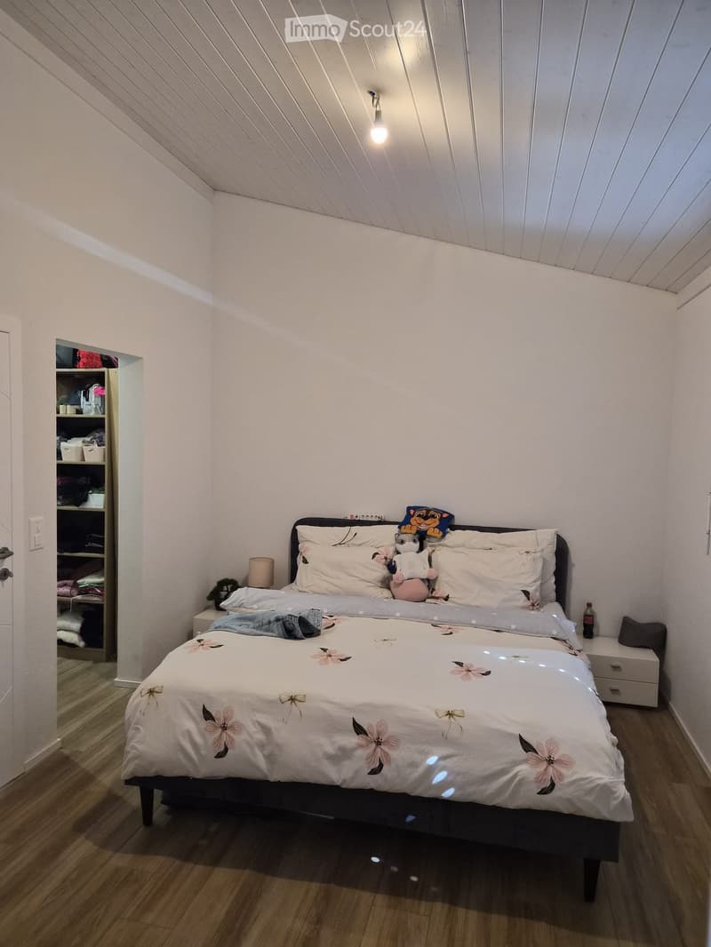 Cozy bedroom with a double bed, white bedding with floral patterns, nightstand with lamp, shelf with folded clothes, and stuffed animals on the bed.