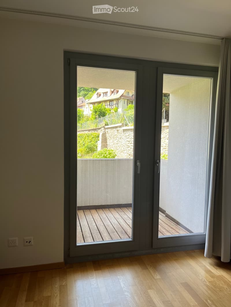 Empty room with wooden flooring, large sliding glass doors leading to a wooden deck, and a view of a garden and neighboring houses.