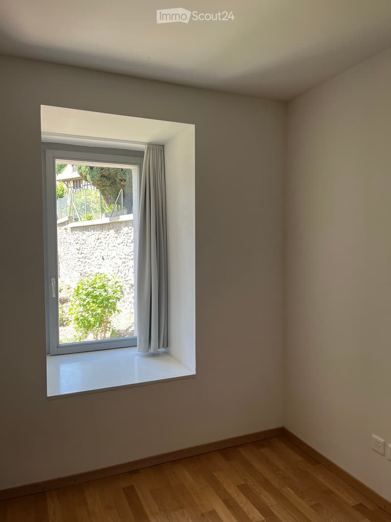 Empty room with wooden flooring, large window with grey curtain, and a view of a stone wall and greenery outside.