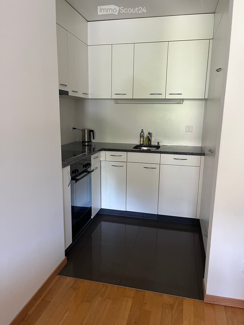 Modern kitchen with white cabinets, black countertops, and a black tiled floor. Features an oven, sink, coffee maker, and a bottle on the counter.