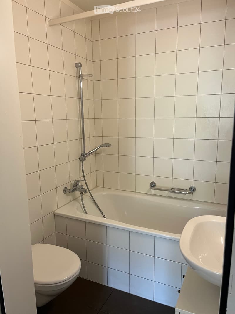 Modern bathroom with a bathtub, toilet, sink, and showerhead. The walls are tiled in light beige, and the floor is dark grey.