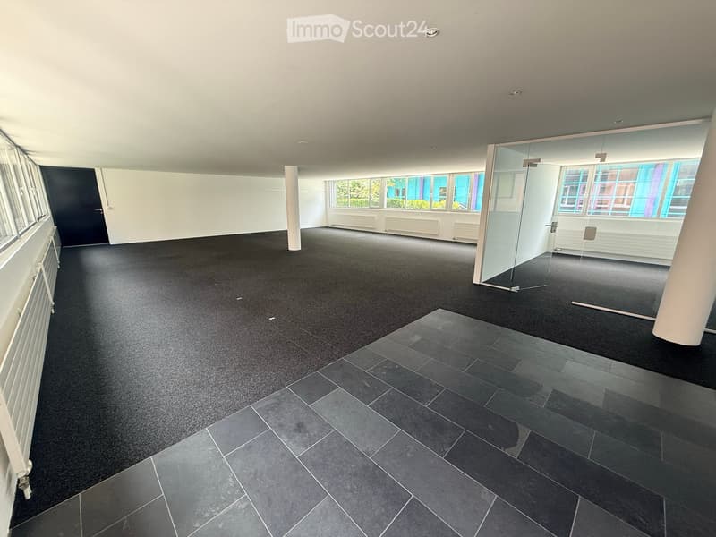 Empty room with dark carpet and tiled section, large windows, glass partition, and white walls.