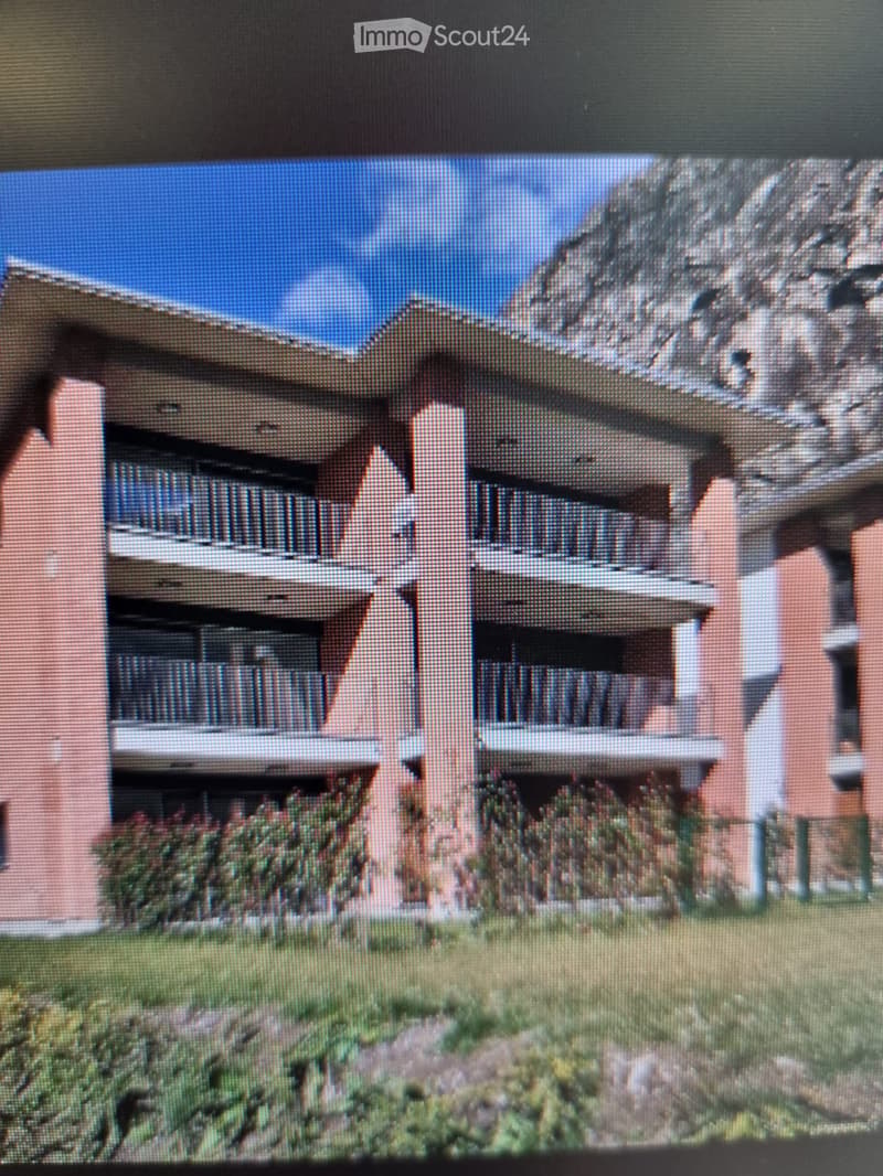 Two-story building with a flat roof, multiple windows, and balconies, surrounded by greenery and a landscaped area.