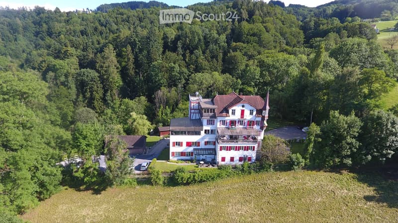 A large, multi-story house with red shutters and a red-tiled roof, surrounded by lush greenery and trees. The house features a balcony and a parking area.