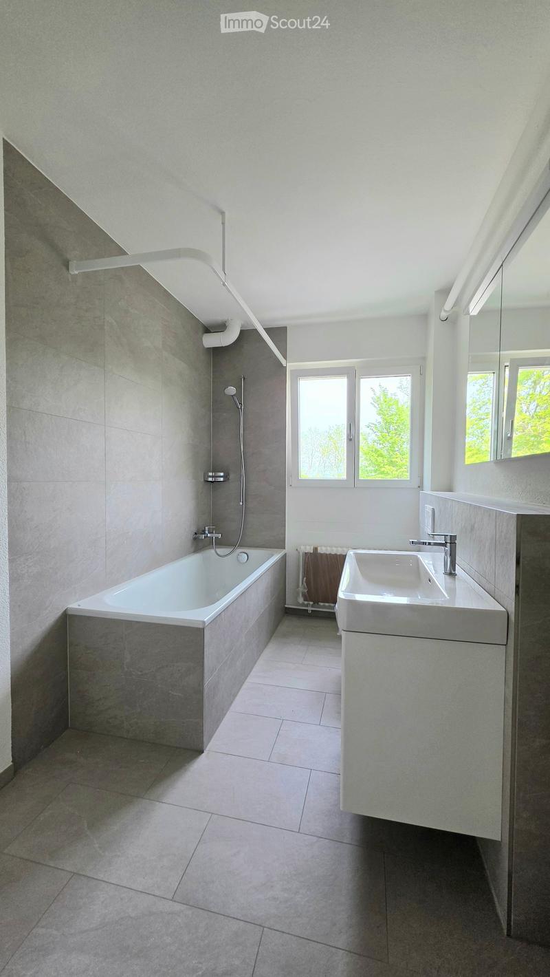 Modern bathroom with a bathtub, sink, and large windows providing natural light. Features include a radiator, a mirror, and sleek fixtures.