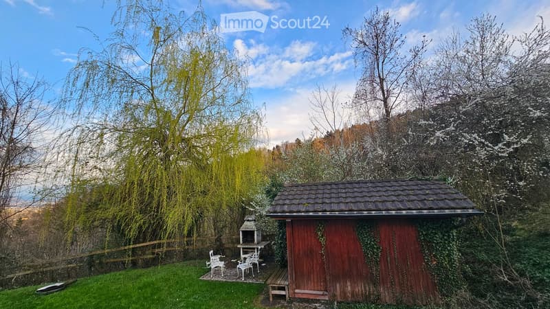 A small wooden shed with a tiled roof is surrounded by greenery and trees. Nearby, there is a small seating area with white chairs and a table, along with a bench and a barbecue.