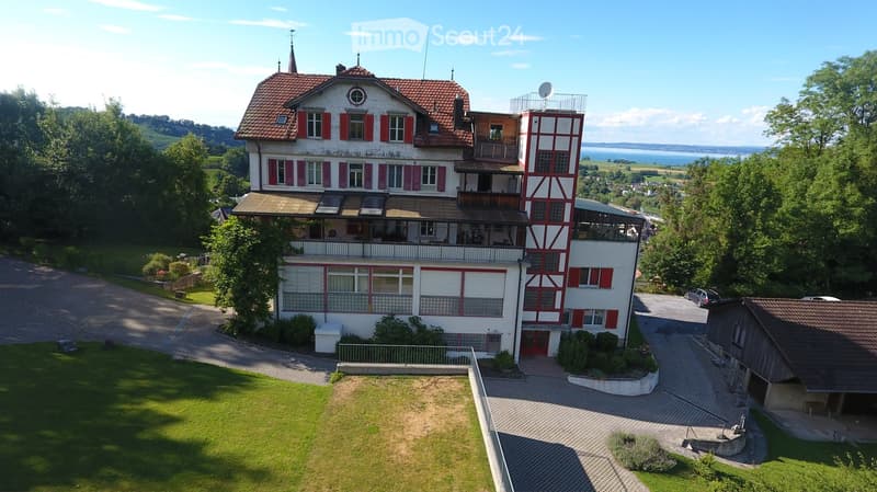 A large, two-story house with red shutters and a red-tiled roof, surrounded by a well-maintained lawn and trees. The house features a balcony and a modern extension with large windows.
