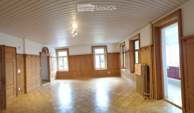 Empty room with wooden panel walls, wooden floor, large windows, and a radiator. No furniture or equipment present.