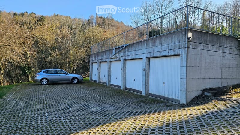 A modern concrete garage with four white doors, a raised ground floor, and a metal railing on the roof. A silver car is parked on a paved driveway.