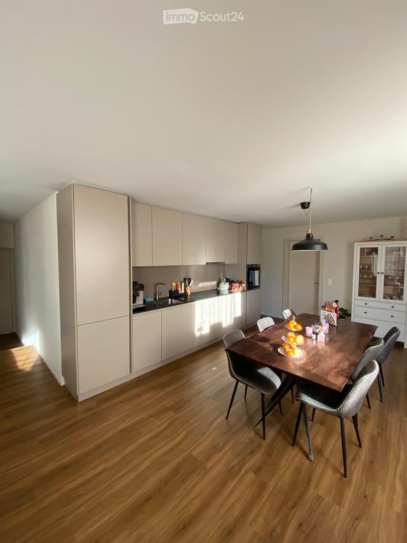 Modern kitchen with light-colored cabinets, a wooden dining table, and six chairs. The kitchen features a sink, faucet, oven, refrigerator, and dishwasher. A cabinet with glass doors is visible, and the floor is made of polished wood.