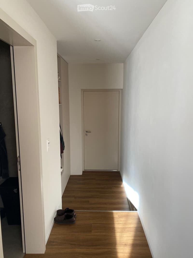 A narrow hallway with wooden flooring, white walls, and a closed door at the end. A pair of shoes is placed near the left wall, and a small shelf with folded clothes is visible in the background.