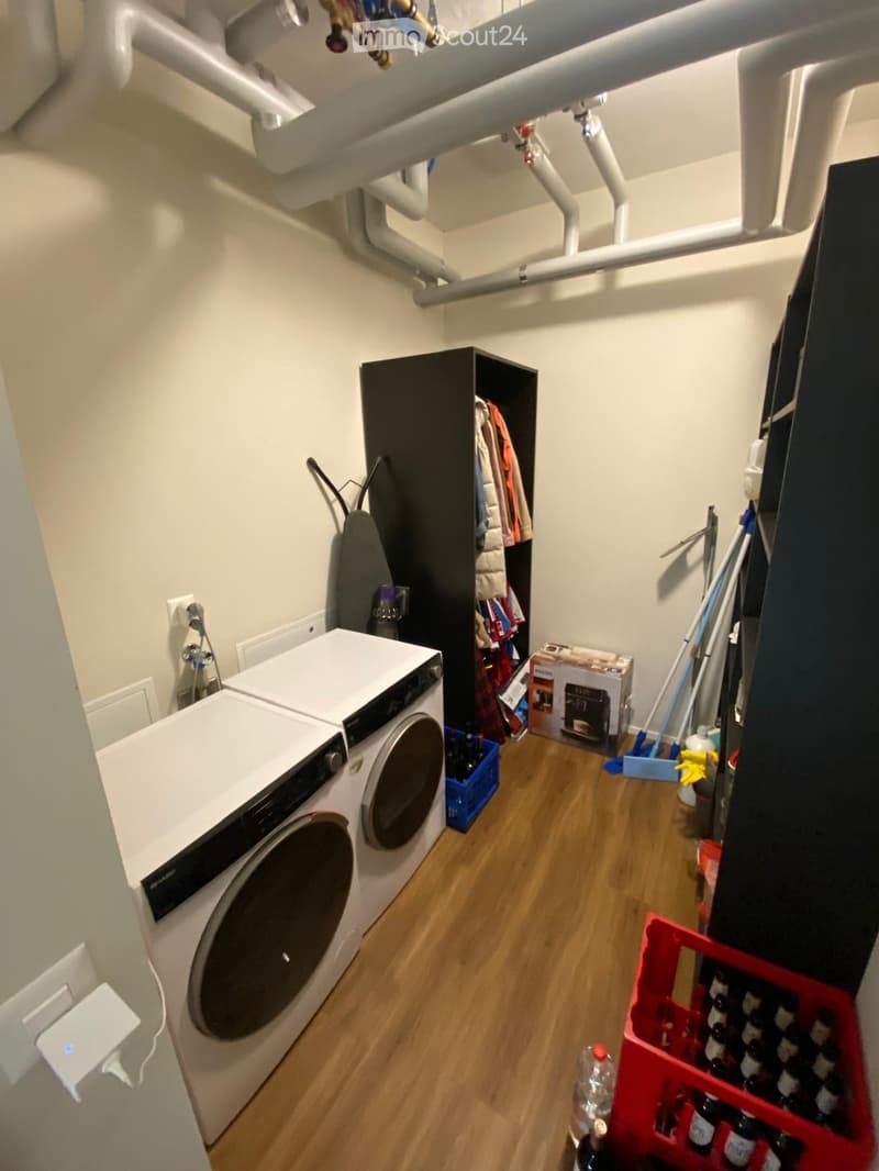 A small utility room with a washing machine, a tall wardrobe, an ironing board, cleaning supplies, and storage boxes.