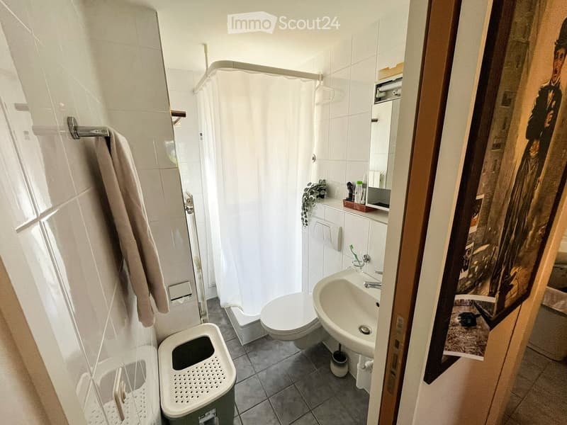 Modern bathroom with white tiles, a shower, toilet, sink, and a mirror. Includes a towel, toothbrush, and a trash bin.