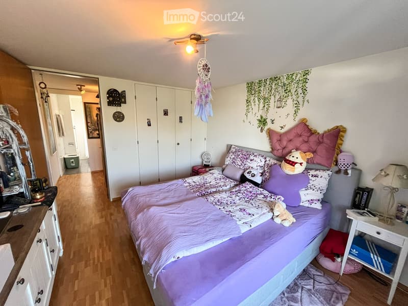 Cozy bedroom with a single bed, colorful pillows, stuffed animals, a nightstand, a mirror, and a wardrobe. The room features wooden flooring and a decorative dreamcatcher hanging from the ceiling.