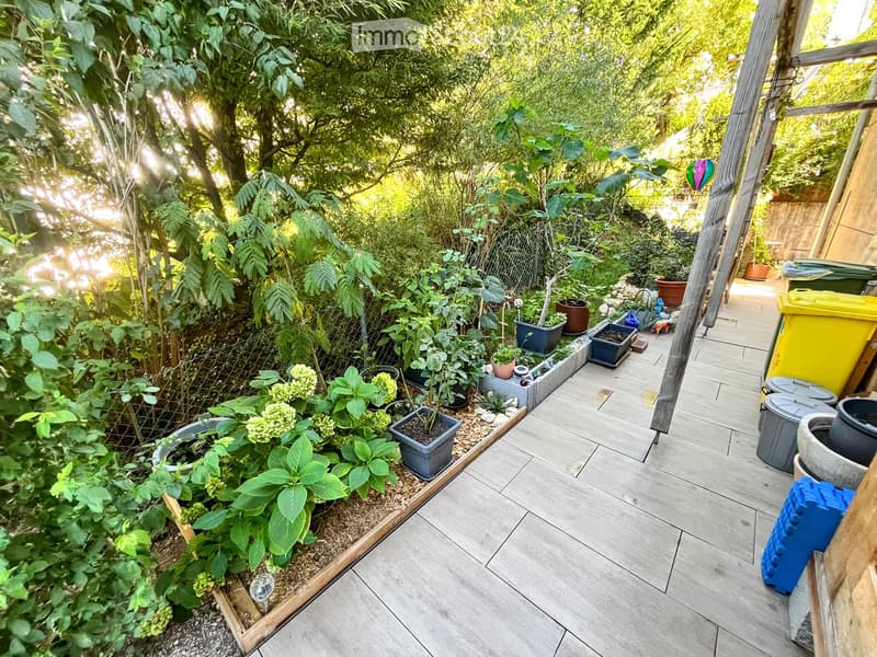 Garden area with wooden decking, potted plants, and lush greenery, including trees and shrubs, with a small raised bed and various containers.