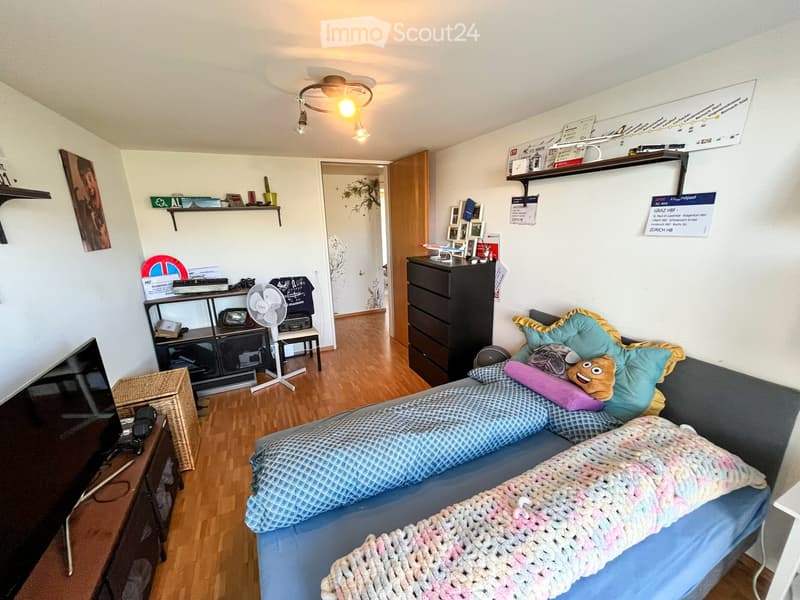A cozy bedroom with a blue sofa, a bed with colorful blankets and pillows, a small table with a fan, shelves with various items, and a ceiling light fixture.