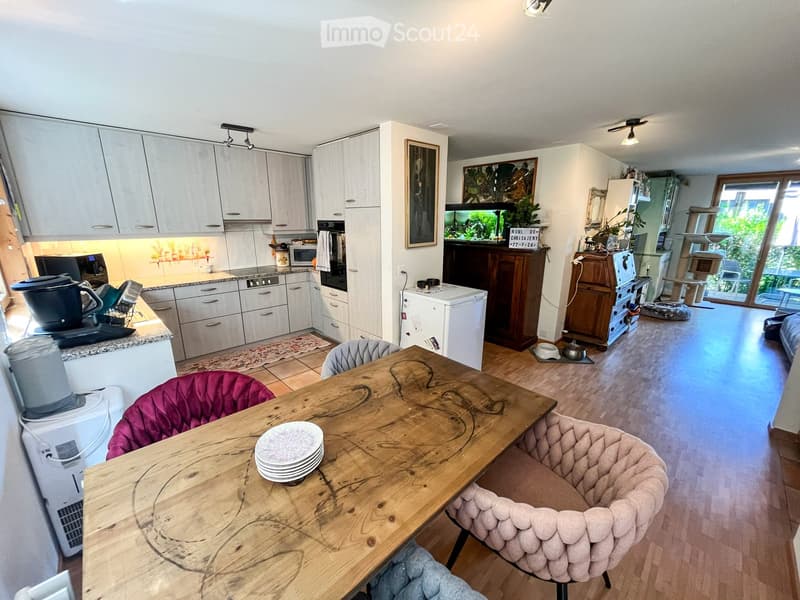 Open-plan kitchen and living area with modern white cabinets, a wooden dining table with chairs, a fish tank, and a cat tree. The kitchen features stainless steel appliances, including a refrigerator, oven, and microwave, and a countertop with various ite