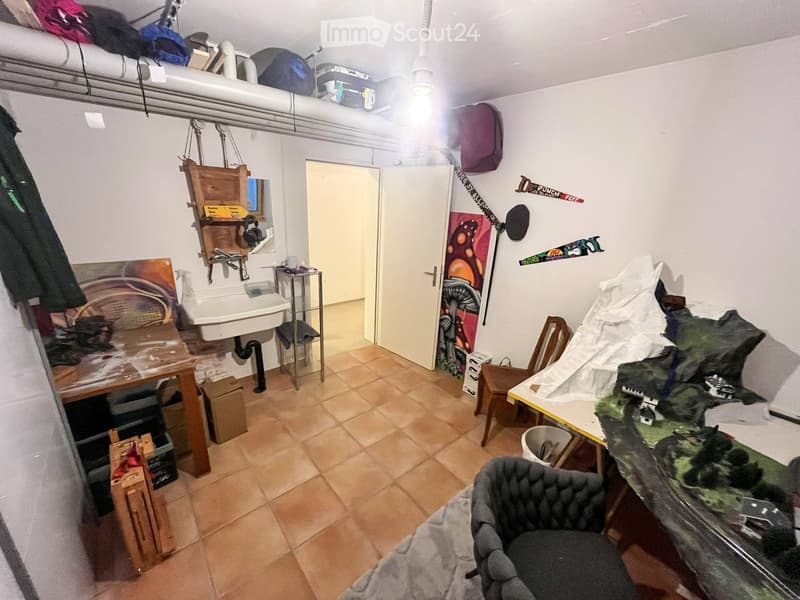 The room is furnished with a chair, table, sink, cabinet, mirror, painting, stool, faucet, and shelf. It has tiled flooring and white walls.
