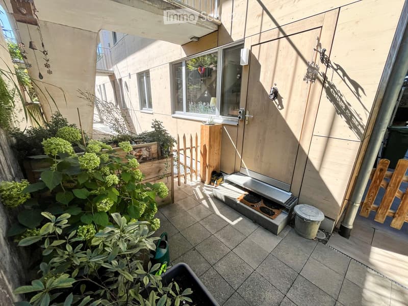 A small outdoor patio area with tiled flooring, potted plants, a wooden fence, and a door leading to the building.