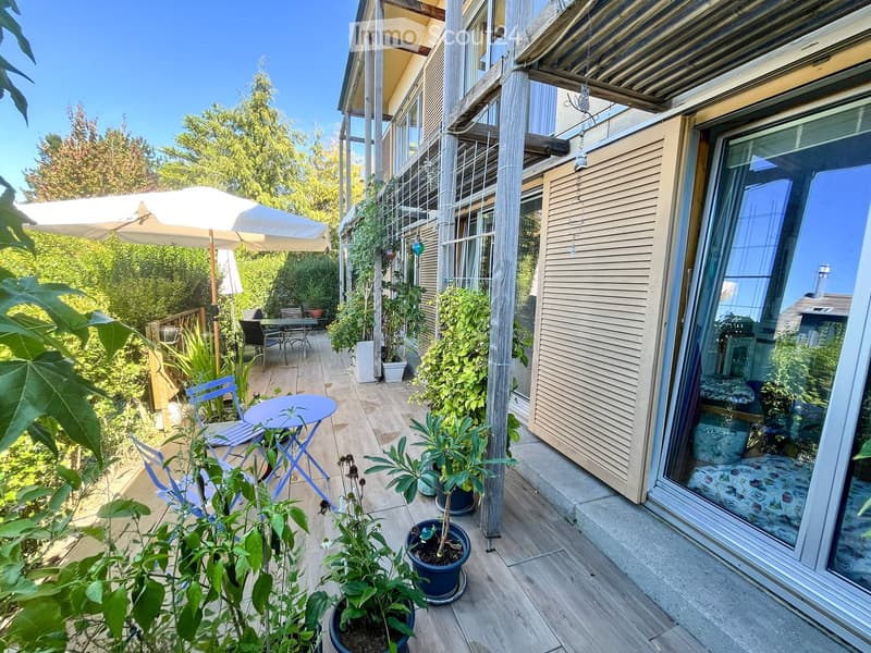 Balcony with wooden decking, potted plants, a small table, chairs, and an umbrella. Large sliding glass door leads to the interior.
