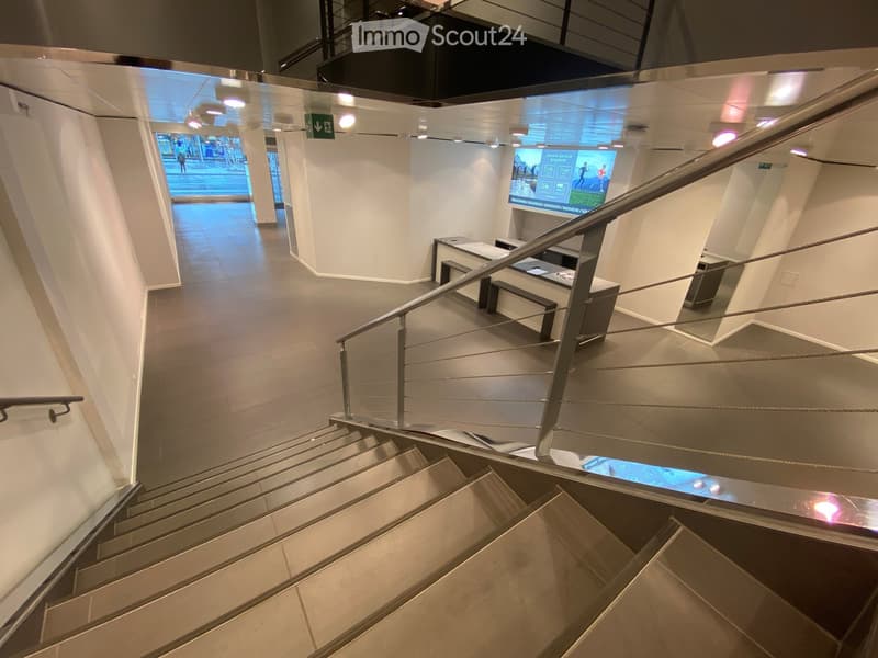Modern hallway with tiled floors, white walls, and a staircase with metal railings. A bench and a digital screen are visible in the background.