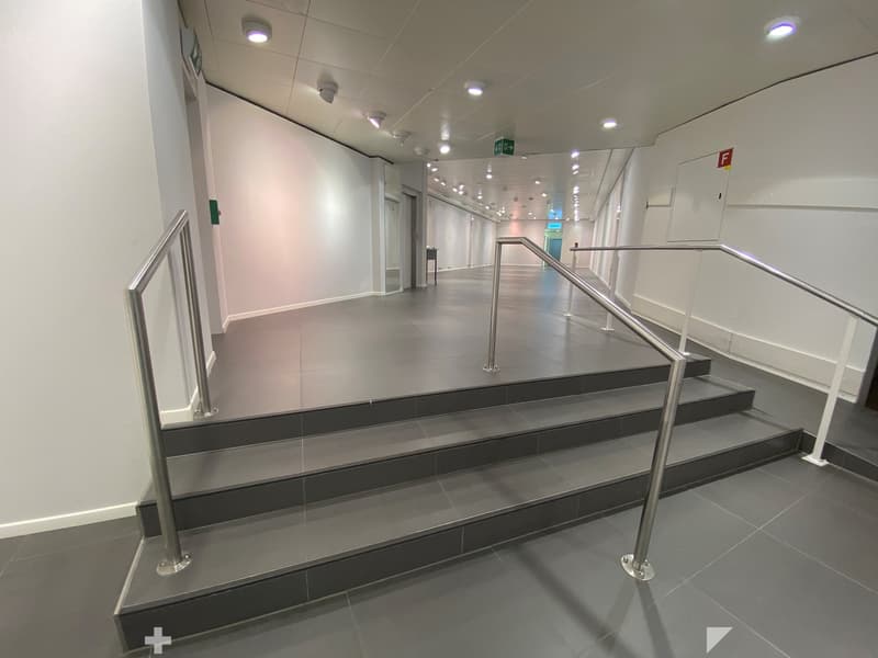 Modern hallway with tiled floors, white walls, and metal railings leading to a staircase. Recessed lighting and emergency exit signs are visible.