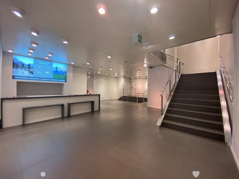 Modern hallway with tiled floor, staircase, reception desk, and digital screen displaying multilingual welcome message.