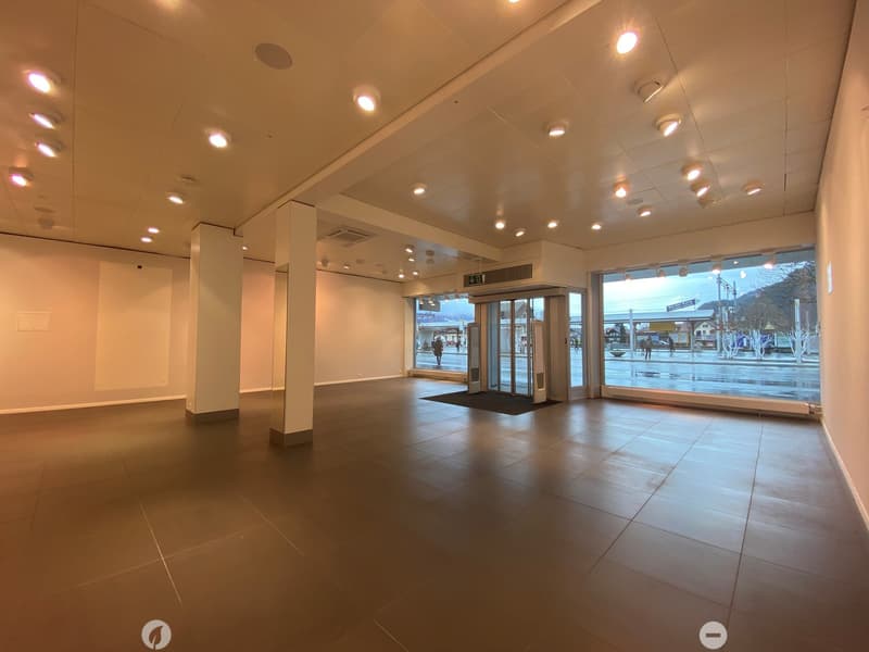 Empty, spacious room with tiled floor, white walls, and large windows overlooking a street. Minimalist design with recessed lighting and a central pillar.