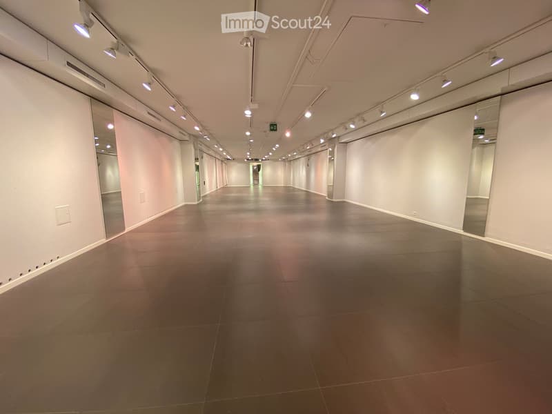 Empty hallway with gray tiled floor, white walls, and recessed lighting. Mirrors on both sides and a green exit sign visible at the far end.