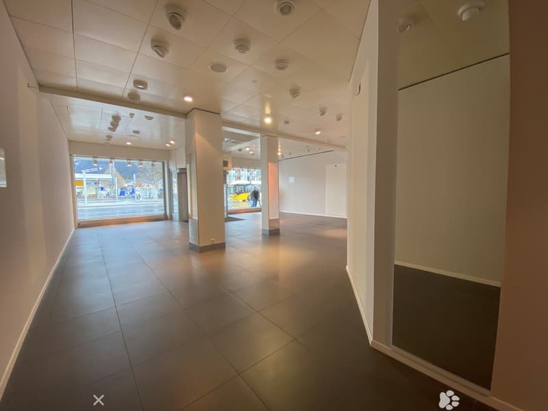 Empty hallway with tiled floors, white walls, and recessed lighting. Features a large window at one end showing a street view.