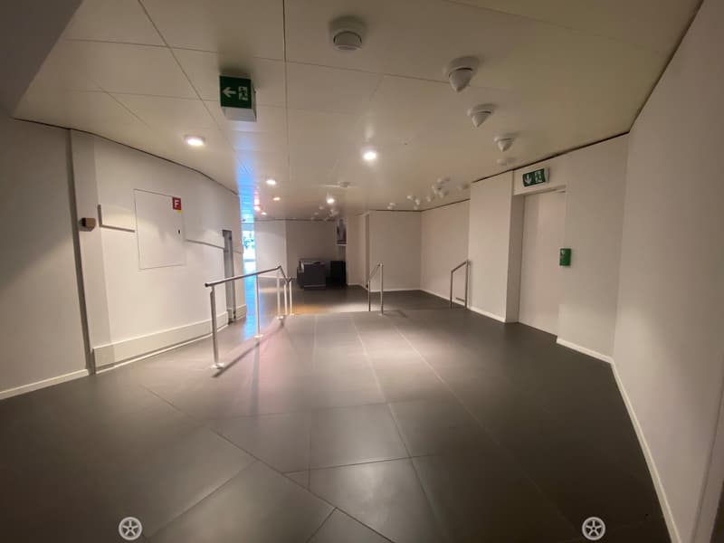 Modern hallway with gray tiled flooring, white walls, and metal railings on both sides. Emergency exit signs and a fire alarm box are visible.