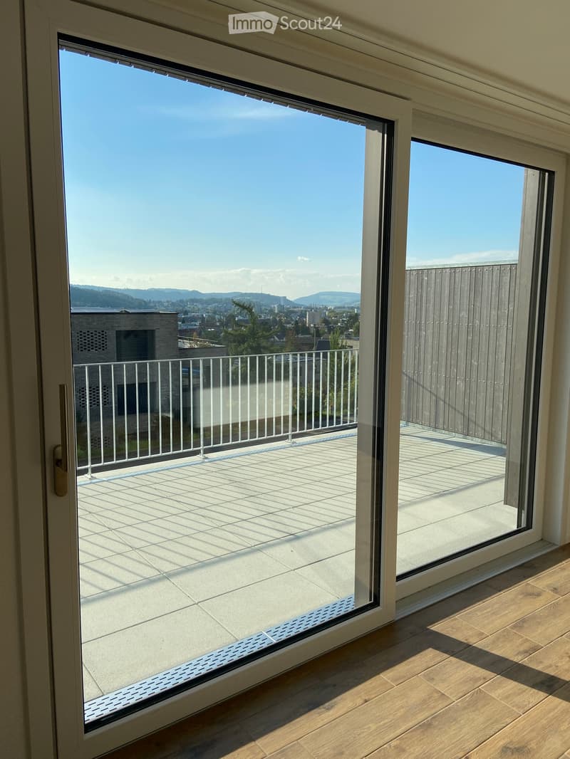 View from inside a room through large glass doors onto a modern balcony with a white railing and tiled floor, overlooking a cityscape with buildings and distant hills.