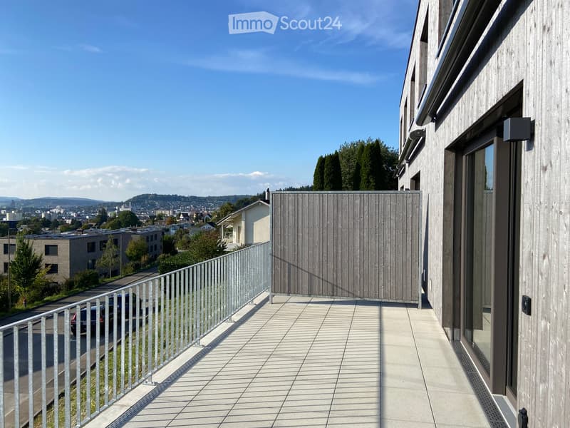 Modern balcony with wooden paneling, metal railing, and tiled flooring, overlooking a cityscape with buildings and trees.