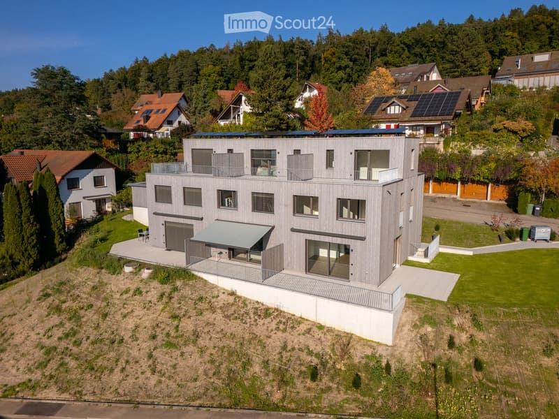 Modern two-story house with grey wooden facade, large windows, solar panels on the roof, garage, and a covered outdoor patio area.