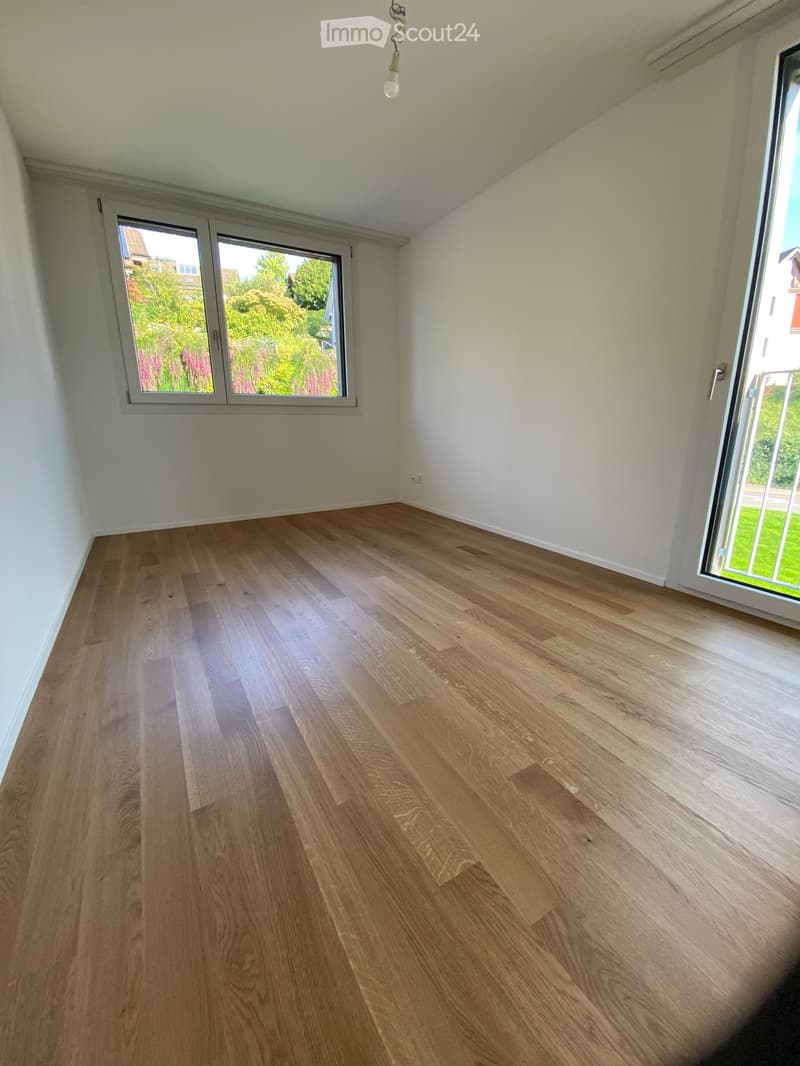 Empty room with light wooden flooring, white walls, and large windows offering a view of greenery outside.