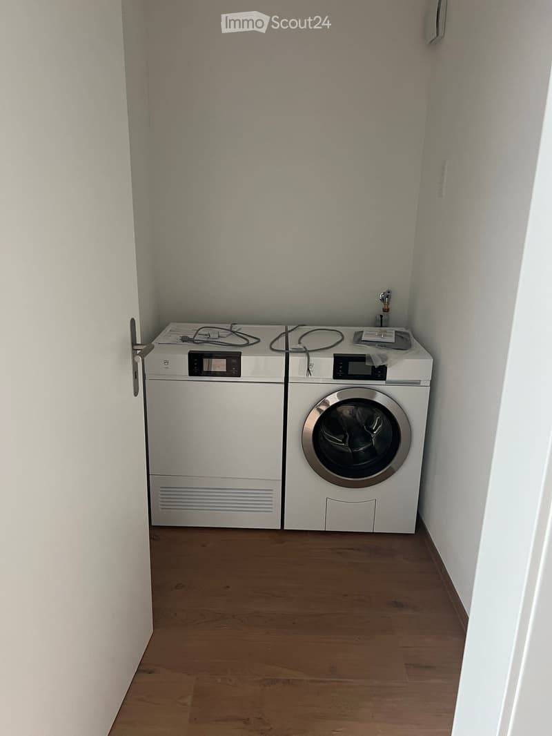 A small room with wooden flooring and white walls, containing a washing machine and a dryer.