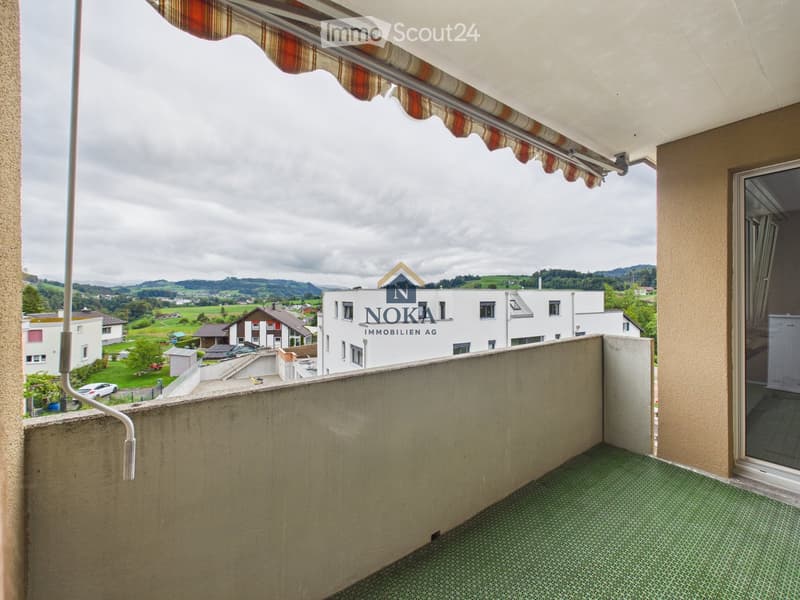 Balcony with green tiled floor, beige walls, and a striped awning extending over the edge. The view shows a residential area with houses, fields, and distant hills.