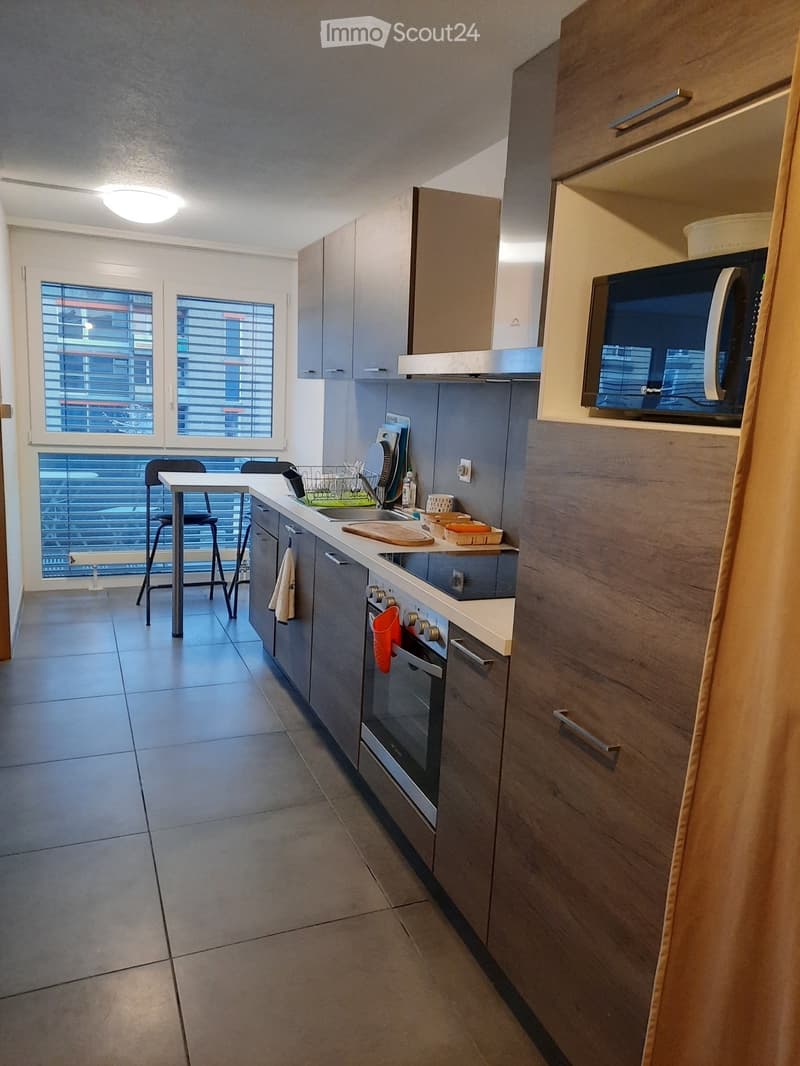 Modern kitchen with grey cabinets, stainless steel appliances, microwave, oven, and a small dining area with a table and stools. The kitchen features a countertop with a dish rack, cutting board, and various small items.