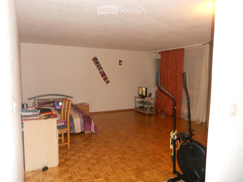 A furnished bedroom with a bed, desk, chair, laptop, TV stand, and exercise bike. The room features wooden flooring and white walls.