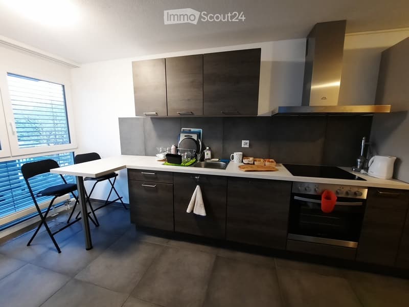 Modern kitchen with grey cabinets, white countertop, built-in oven, and a small dining area with a table and chairs. The kitchen features a sink, dish rack, and various appliances including an oven and a refrigerator.