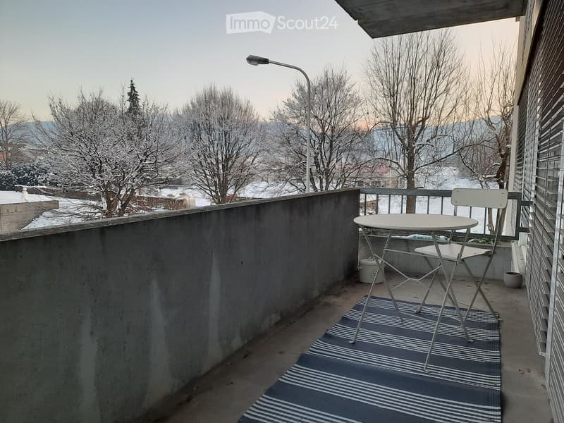 Balcony with a small table and chair, overlooking a snowy landscape with trees and distant mountains.