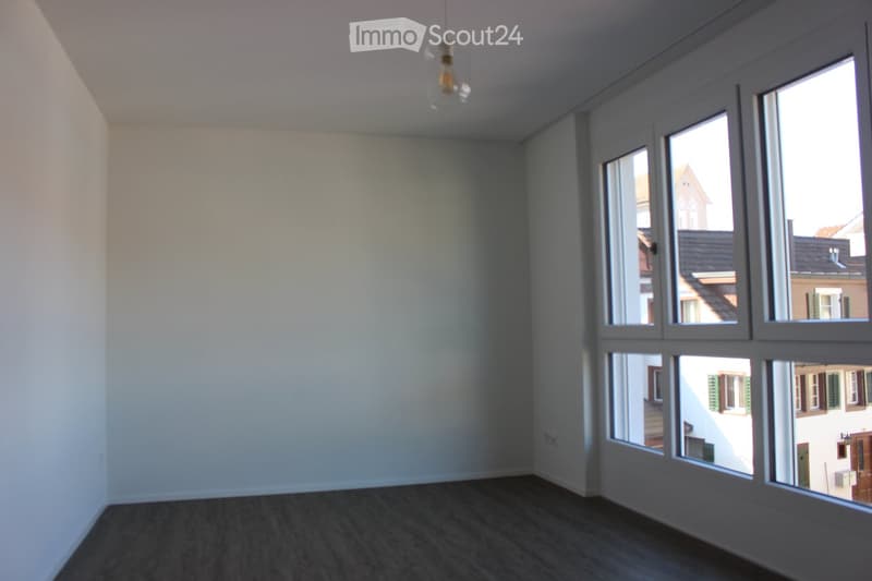 Empty room with light-colored walls, dark wooden flooring, and large windows offering a view of neighboring buildings.