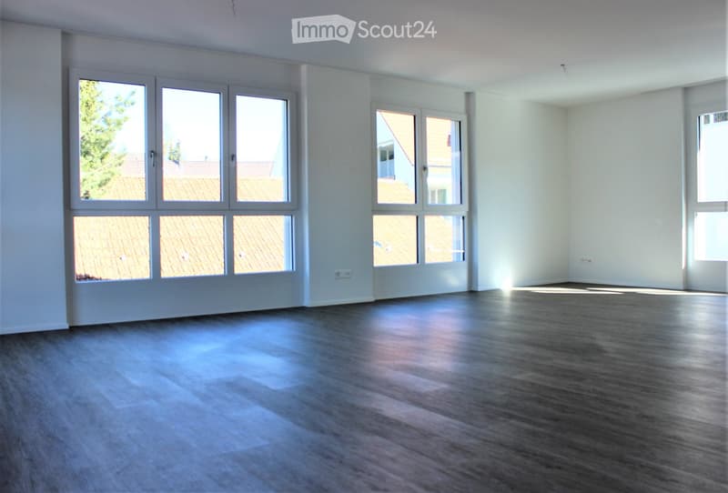 Empty room with light-colored walls and wooden flooring, featuring large windows with views of rooftops and trees.