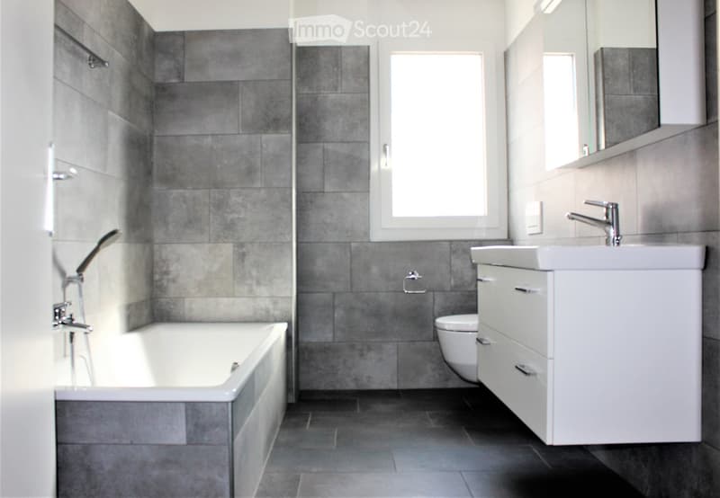 Modern bathroom with grey tiled walls, a white bathtub, a white sink with a chrome faucet, a toilet, and a mirrored cabinet.
