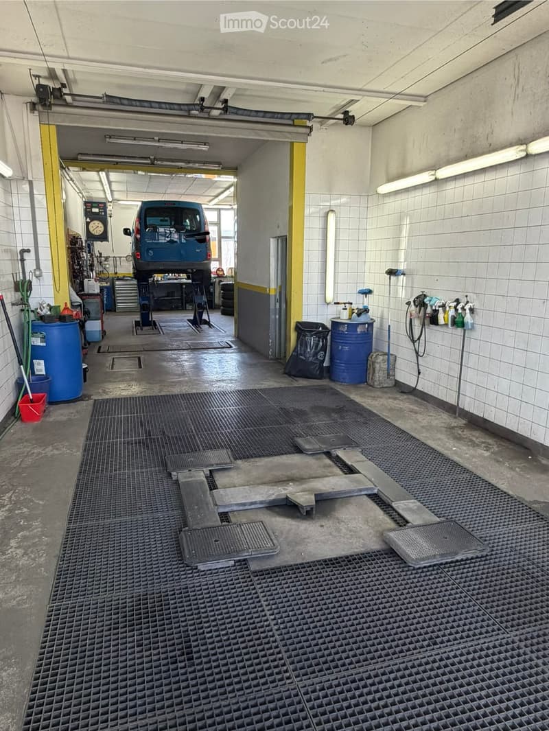 A car service area with a raised car on a lift, surrounded by tools, cleaning supplies, and a concrete floor with drainage grates.