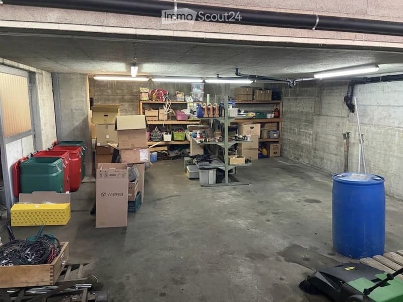 A spacious underground garage with concrete floors and walls, filled with various storage items including cardboard boxes, shelves, storage bins, barrels, and tools.