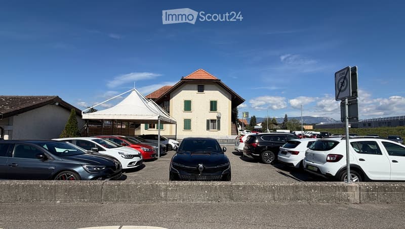 A multi-story building with a red-tiled roof and green shutters, surrounded by a parking lot with several parked cars under a clear blue sky.