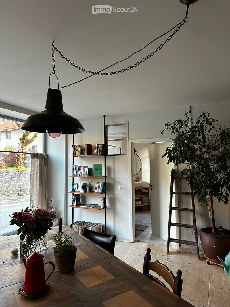 Bright living space with wooden flooring, large window, bookshelf filled with books, dining table with chairs, potted plants, and a ladder leaning against the wall.
