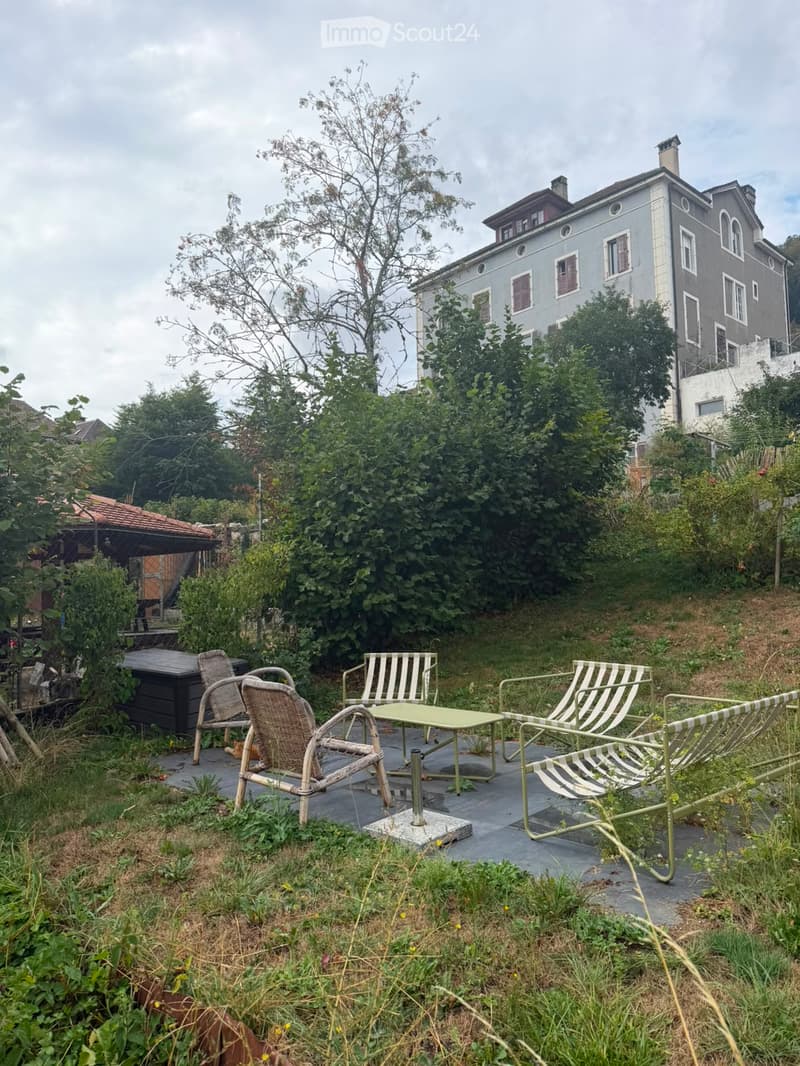 Outdoor seating area with a table, chairs, and lounge chairs, surrounded by greenery and a large building in the background.